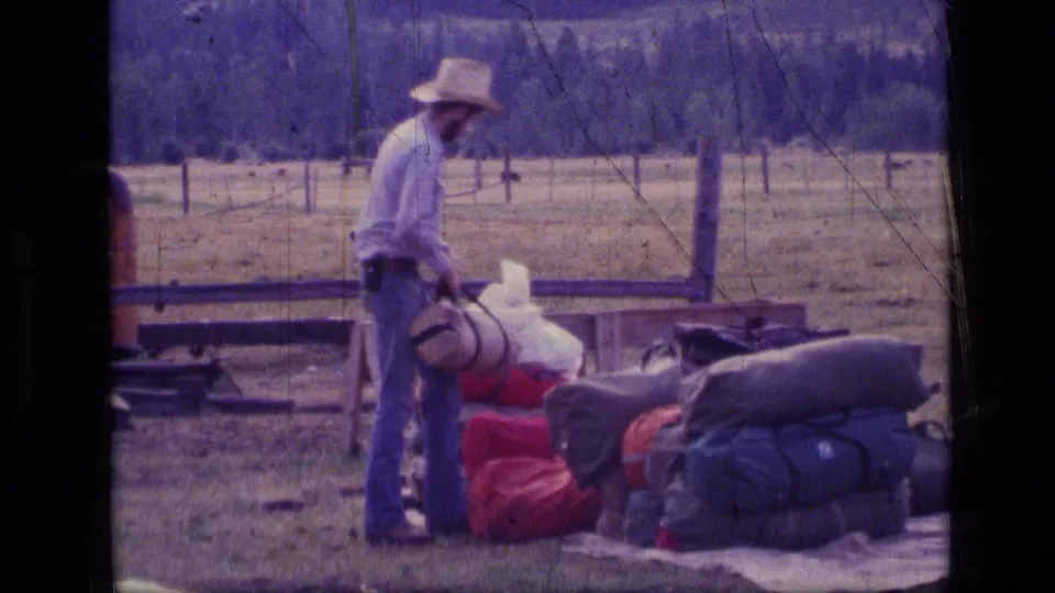 1973 Montana 8mm frame of a man in a cowboy hat handling camping gear beside duffel bags, tarps, and a wooden fence.