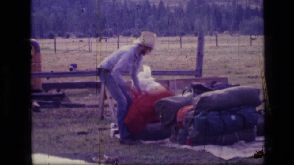 1973 Montana 8mm frame showing a man bending to organize red duffel bags and sleeping rolls in a fenced wilderness field.