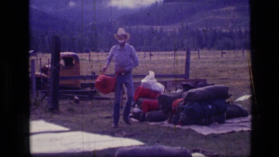 1973 Montana 8mm frame of a man carrying a red duffel near sleeping rolls, tarps, a fence, and an older truck in the background.