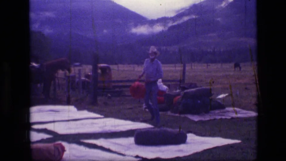 1973 Montana 8mm frame of a man standing near packed camping gear, a horse, an older truck, fence posts, and distant mountains.