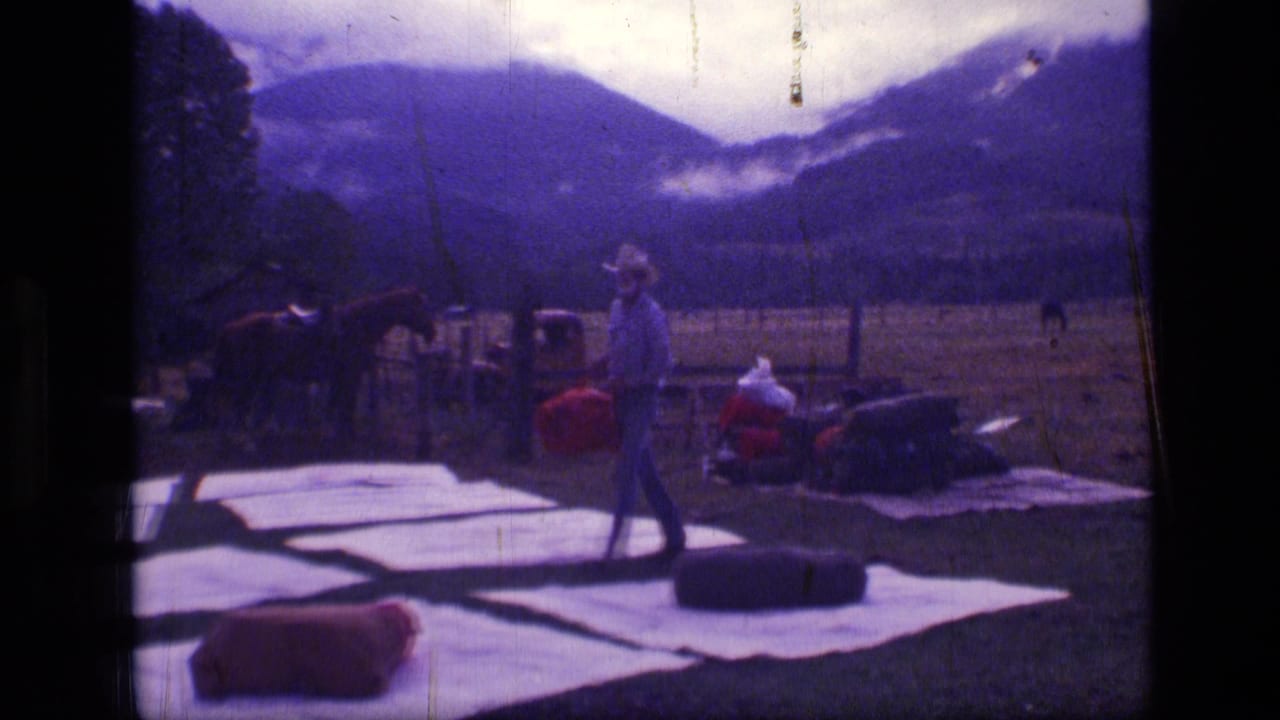 Cowboy carrying a red pack across white tarps in a mountain field with horses nearby.