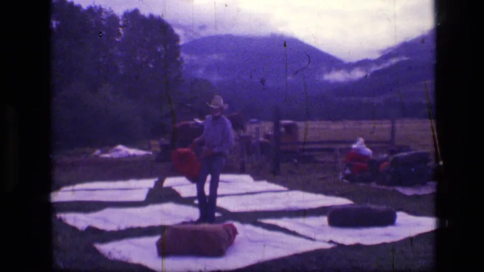 Cowboy in a hat stands among white tarps and red supplies in a likely Scapegoat Wilderness mountain field with horses nearby.