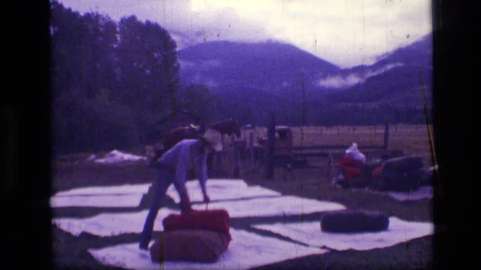 1973 8mm wilderness frame showing a cowboy bending to adjust a red roll on tarps as horses wait in a grassy mountain setting.