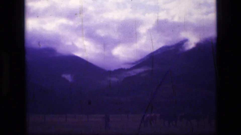Distant 1973 Montana mountain-pasture frame with horses near the lower edge and a dramatic clouded sky above dark slopes.