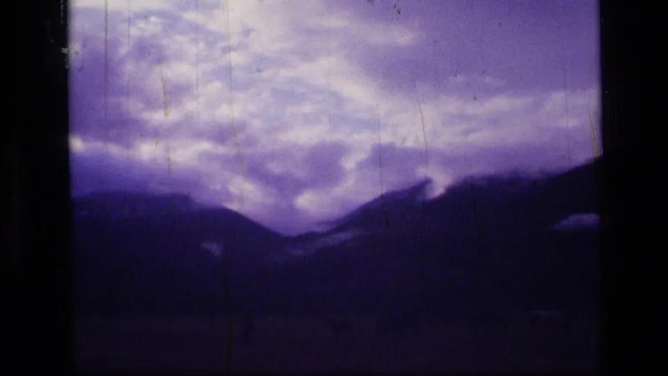 1973 Montana wilderness home-movie frame showing a cloudy sky above a dark mountain slope with heavy 8mm color shift and dust marks.