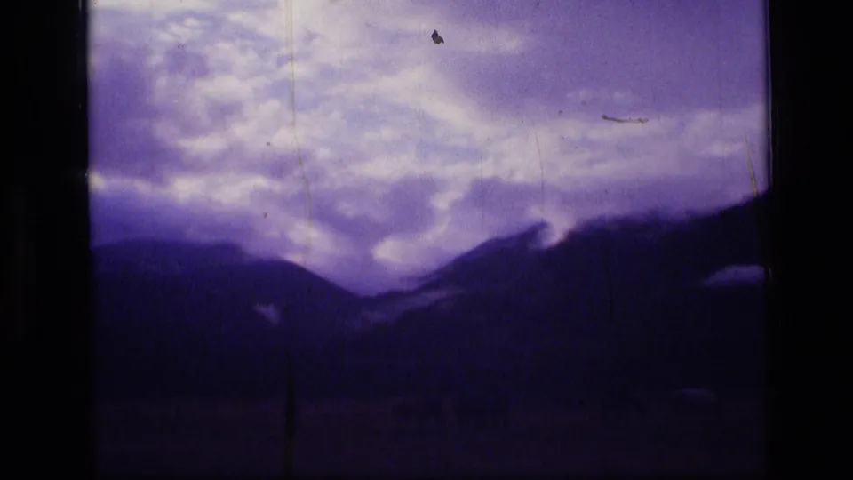 1973 8mm wilderness frame with layered clouds, shadowed Montana mountain ridges, soft focus, and visible archival film scratches.