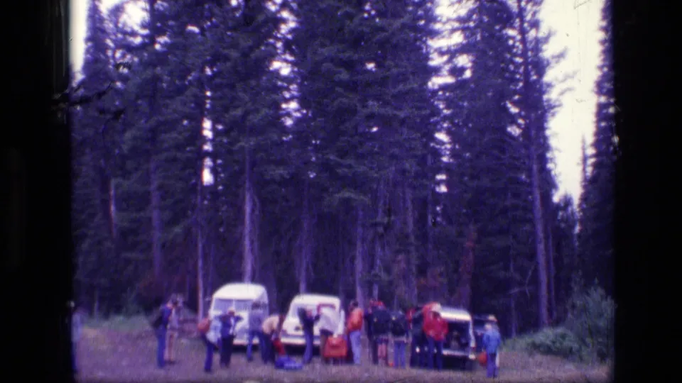 Group of campers in a Montana forest clearing, standing near vintage vehicles among tall conifers in 1973 home-movie footage.