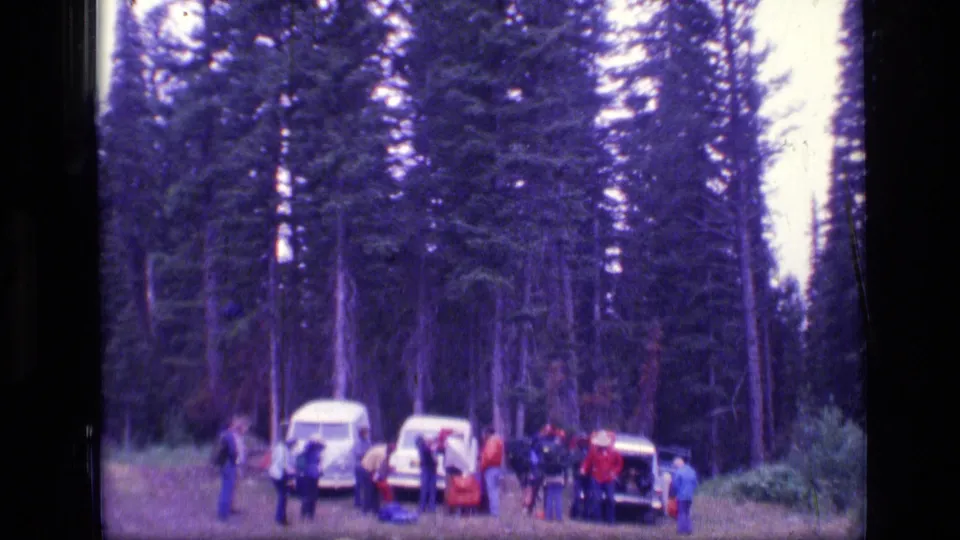 1973 Montana wilderness home-movie frame with a group organizing near a white van and parked cars beneath tall evergreens.