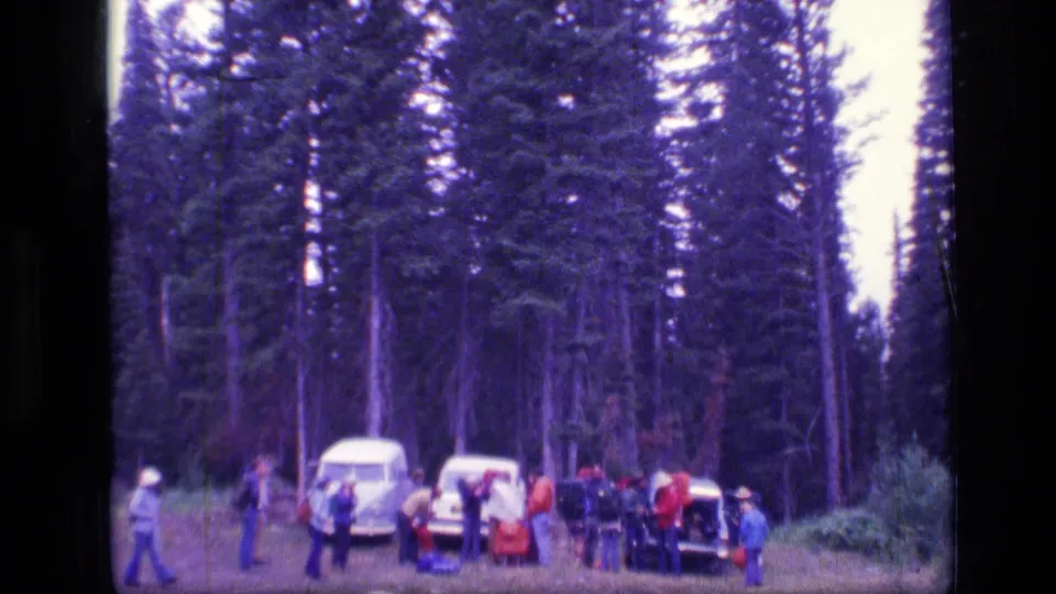 Super 8 frame from the 1973 Montana trip showing adults moving around vehicles and supplies in a forest staging area.