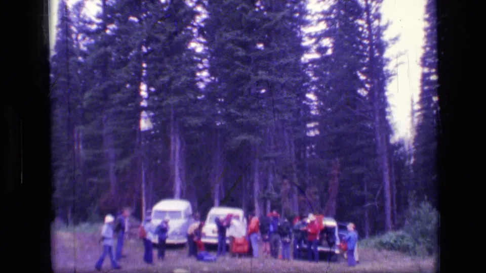 1973 Scapegoat Wilderness home-movie frame with people, vehicles, and gear spread across a conifer-lined clearing.