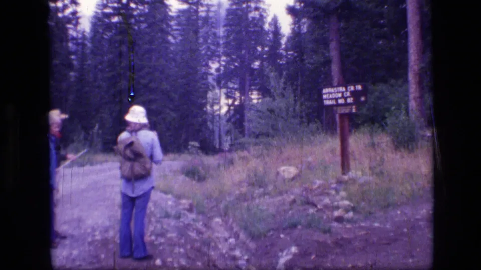 1973 8mm view in Scapegoat Wilderness, Montana, with hikers near a wooden Arrastre Cr Trail sign in forest.