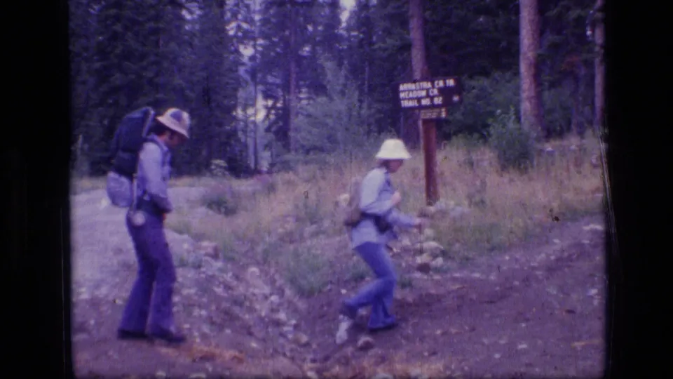 Two hikers separate at the forest junction, with one stepping across rocks and another moving past the wooden sign.