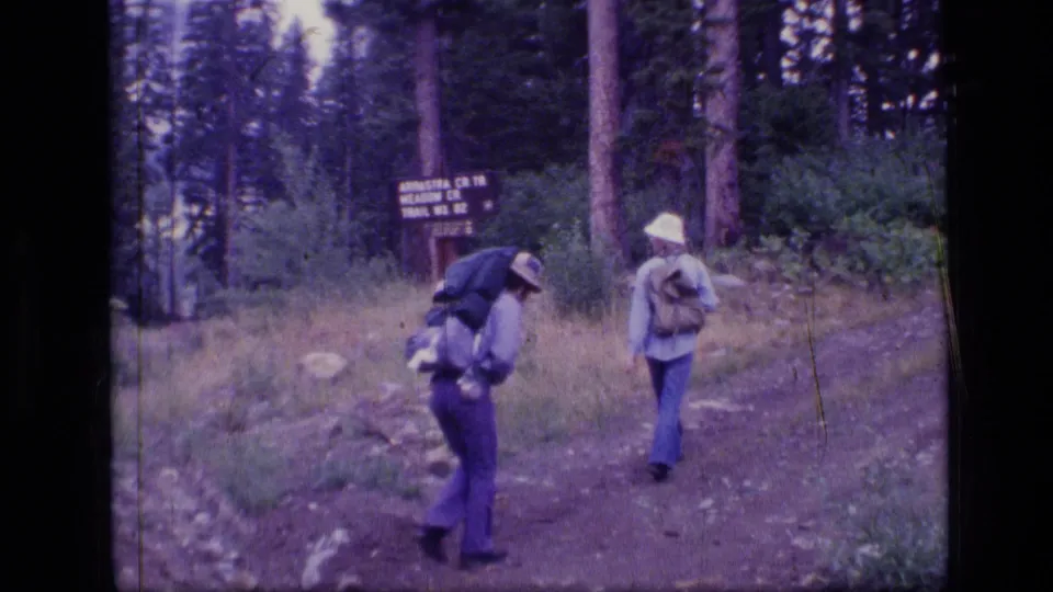 Backpacked hikers move along a forest trail near a sign in 1973 Scapegoat Wilderness footage.