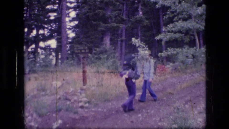 1973 Scapegoat Wilderness home-movie frame with two backpackers near a trail sign among trees and grass.