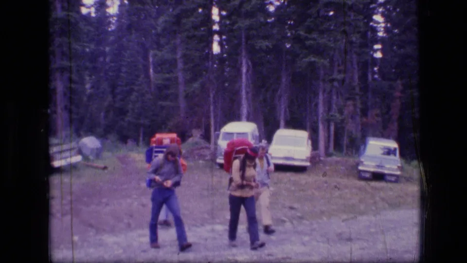 1973 Montana 8mm frame showing backpackers near a forest clearing with several parked 1970s cars behind them.