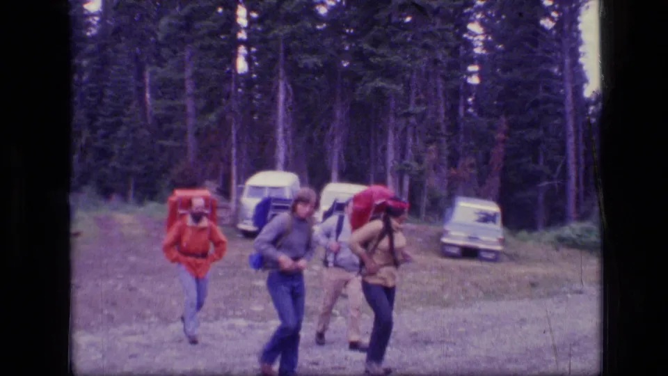 1973 Montana home-movie frame of three backpackers crossing a forest clearing near several parked cars.