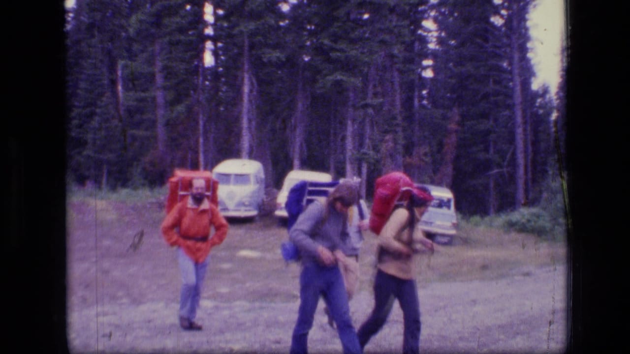 Three adult hikers with large packs walk near parked vehicles at a forest trailhead.
