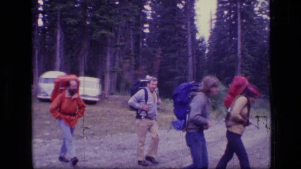 Four adult hikers with external-frame backpacks move through a Montana forest trailhead area in the 1973 home-movie clip.