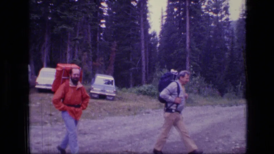 Two backpackers from the 1973 Scapegoat Wilderness footage pass parked vehicles near a dense conifer stand.