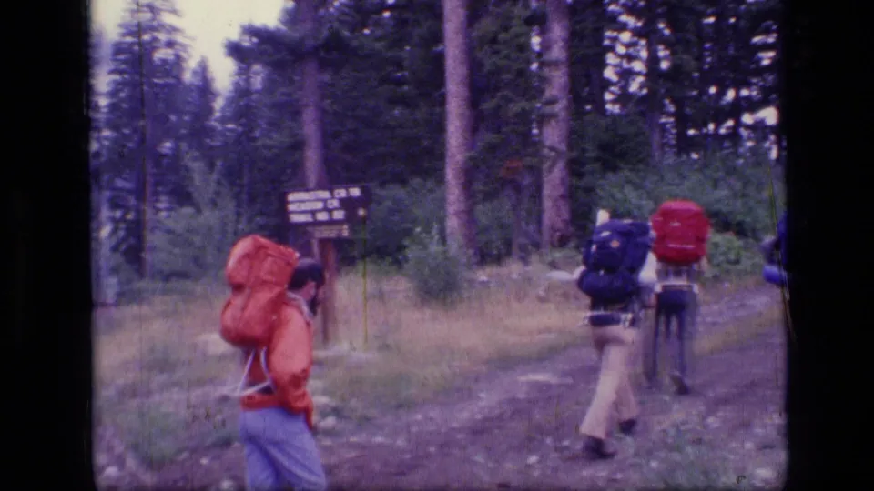 Backpackers walk away into the Scapegoat Wilderness area near a wooden trail sign in 1973 8mm footage.