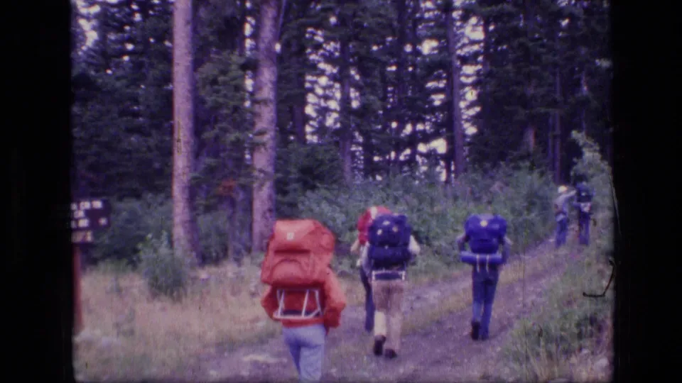 Four hikers with external-frame backpacks continue along a wooded trail in 1970s Montana wilderness footage.