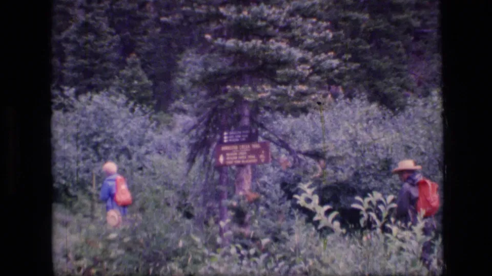 Two backpackers are visible near the trail sign, with one entering from the left and another partly seen at the right edge.