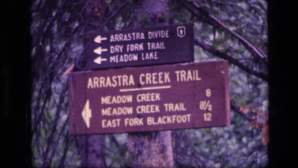 Tight 1973 8mm frame of wooden trail signs for Arrastra Divide, Dry Fork Trail, Meadow Lake, and Arrastra Creek Trail in Montana forest.