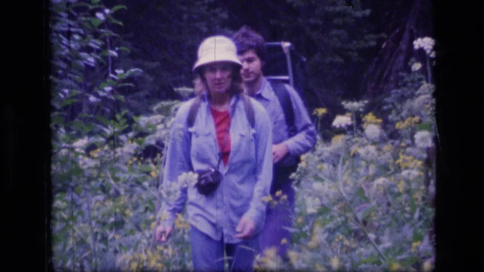 In 1973 Scapegoat Wilderness footage, a woman in a white bucket hat and light jacket walks toward the camera with another hiker behind her.