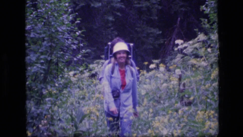 A smiling hiker in a white bucket hat moves through dense Scapegoat Wilderness vegetation, with camera gear visible at her side.