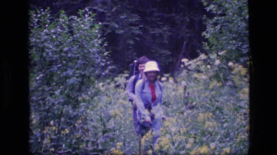 Two adult hikers proceed through a brushy Montana forest trail, the lead hiker wearing a white hat and carrying a camera.