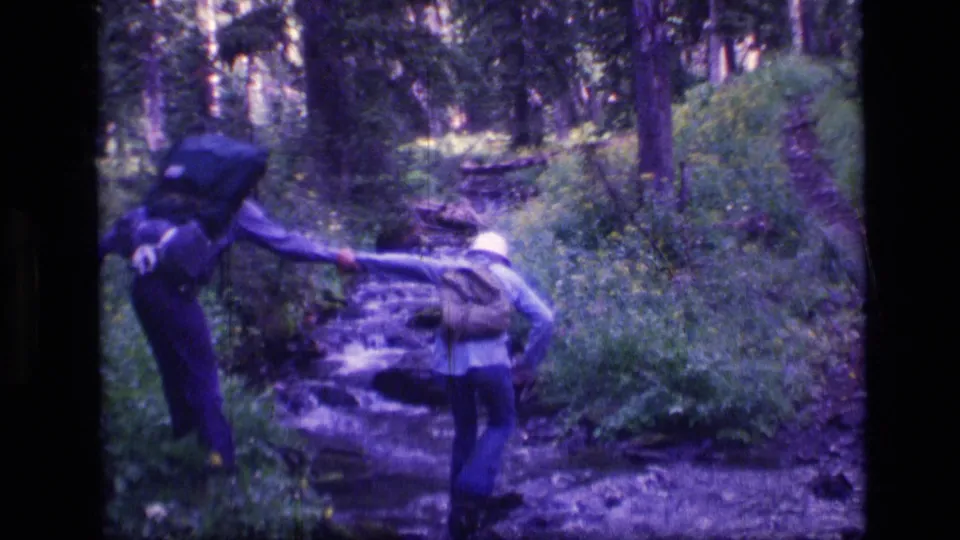 1973 Montana wilderness hiking frame showing one adult assisting another across a narrow forest stream.