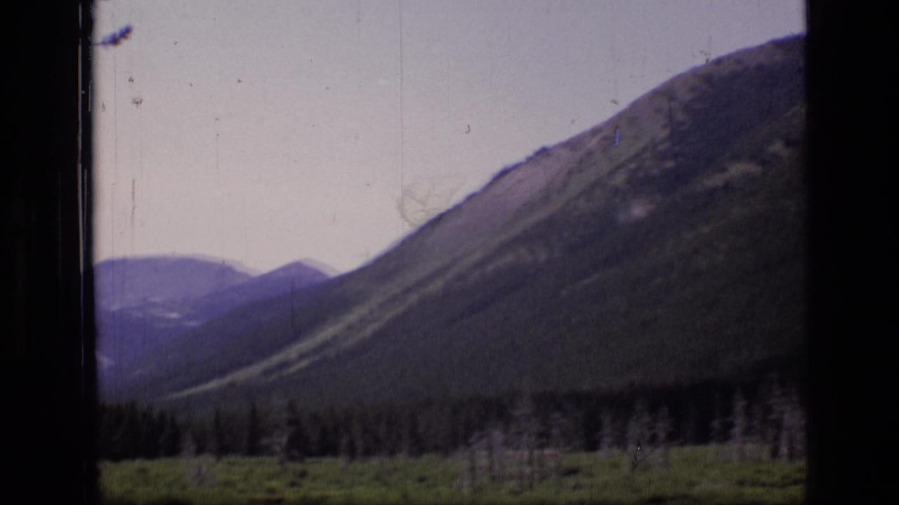 1973 8mm frame showing layered Montana mountain slopes, distant peaks, and a dark forest line under a pale sky.