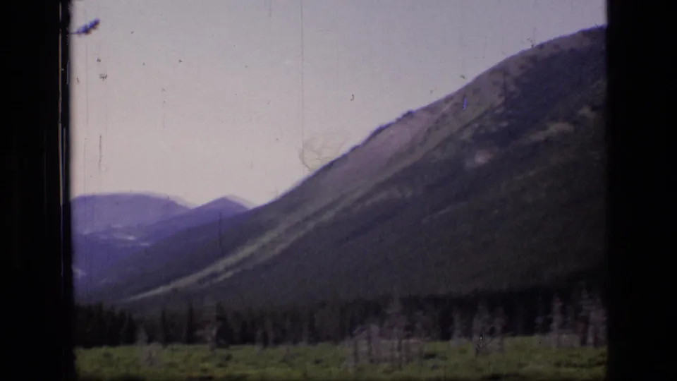 1973 8mm frame showing layered Montana mountain slopes, distant peaks, and a dark forest line under a pale sky.