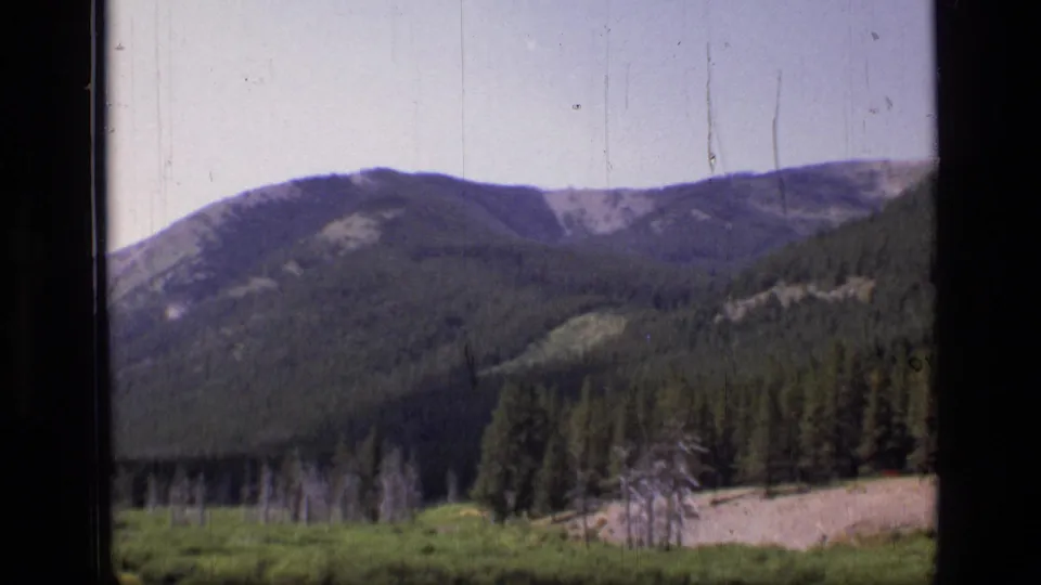 1973 8mm frame of Scapegoat Wilderness terrain with a mountain ridge, evergreen trees, meadow, and pale ground at the edge.