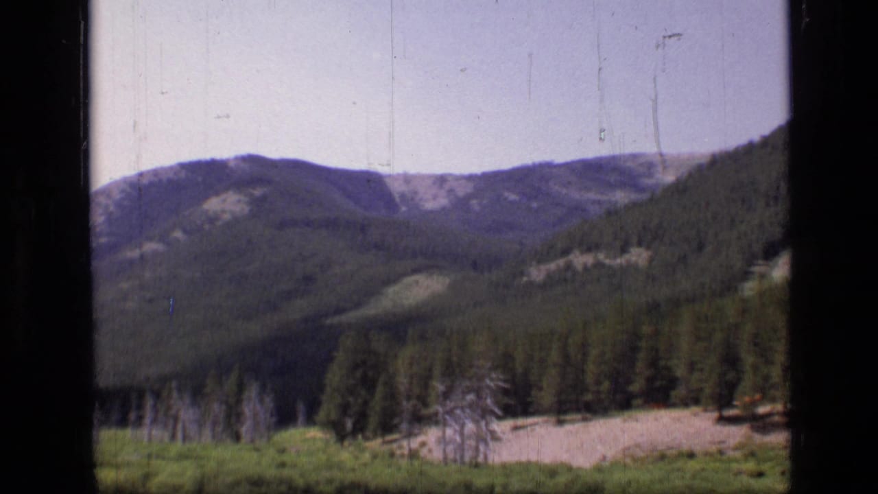 1973 8mm Montana wilderness view of evergreen hills, a pale meadow, and distant ridgelines from a moving vehicle.