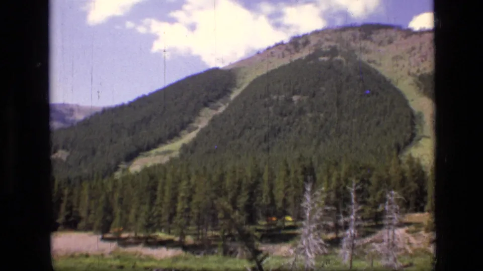 1973 Montana wilderness frame showing a rounded forested mountain, meadow foreground, bare trees, and scattered clouds.