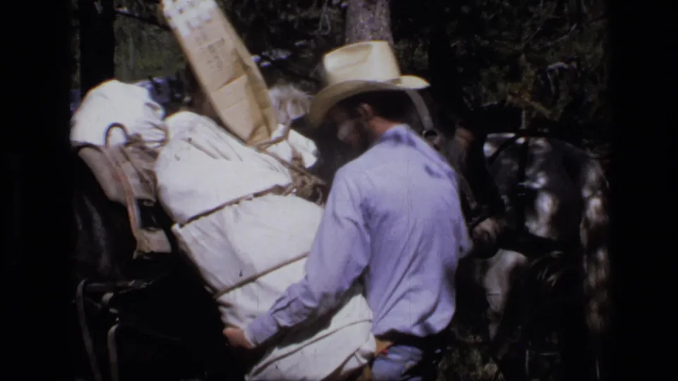 In 1973 Scapegoat Wilderness footage, a man in a cowboy hat handles a white supply sack beside a black horse under trees.