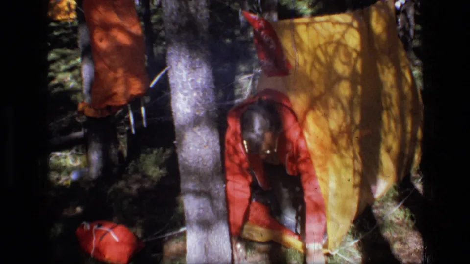 A camper in a red jacket works low beside a yellow tarp shelter and scattered gear in a forest campsite.