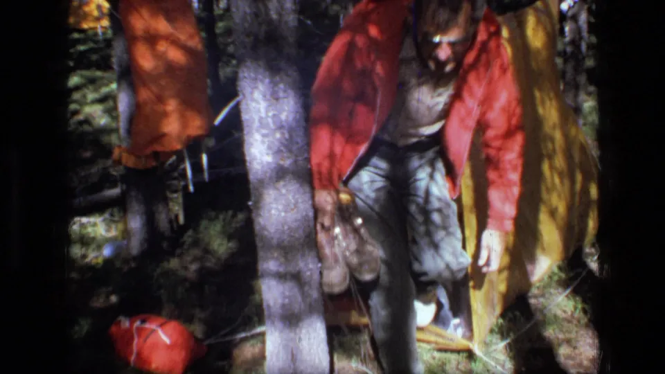 A camper in a red jacket stands near the yellow tarp shelter holding camp items in the Montana wilderness setting.