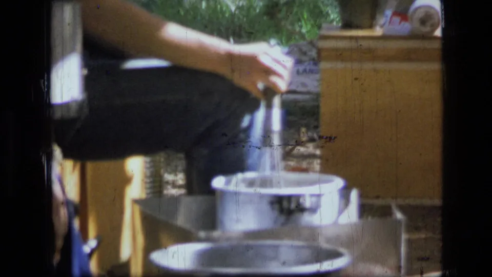 Young woman at a Montana wilderness campsite pours liquid into a metal cooking pot beside a wooden camp chest.