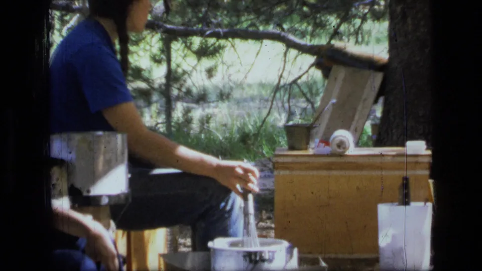 Young woman in a blue shirt sits at a 1973 Montana campsite beside cooking pots, a wooden chest, and forest branches.