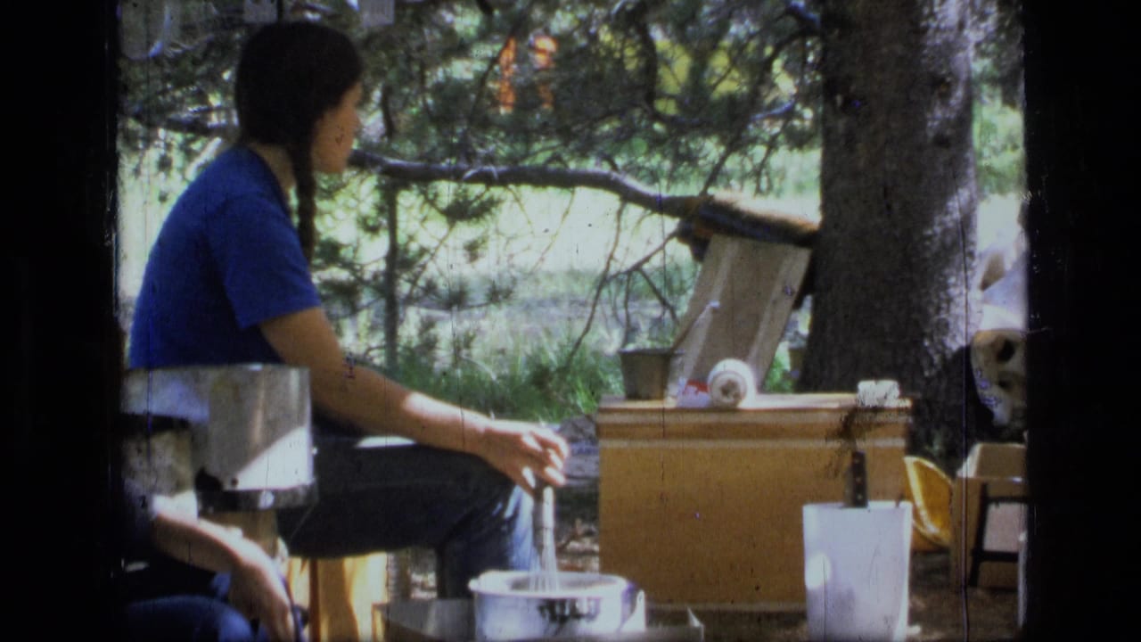 1973 Montana wilderness home-movie frame of a seated camper in blue near pots, boxes, and a shaded forest camp setup.
