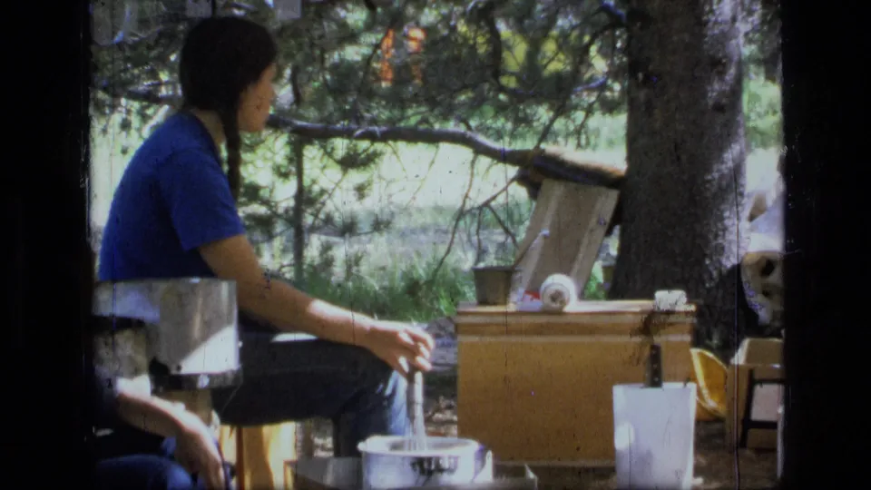 1973 Montana wilderness home-movie frame of a seated camper in blue near pots, boxes, and a shaded forest camp setup.