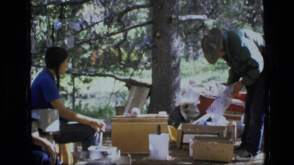 1973 Montana camping frame with one camper seated by cookware while another bends over boxes and fabric under pine trees.