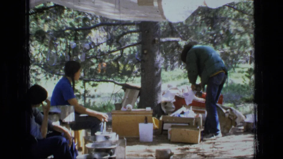 1973 Montana home-movie frame showing campers around a forest campsite with cookware, storage boxes, and a tarp overhead.