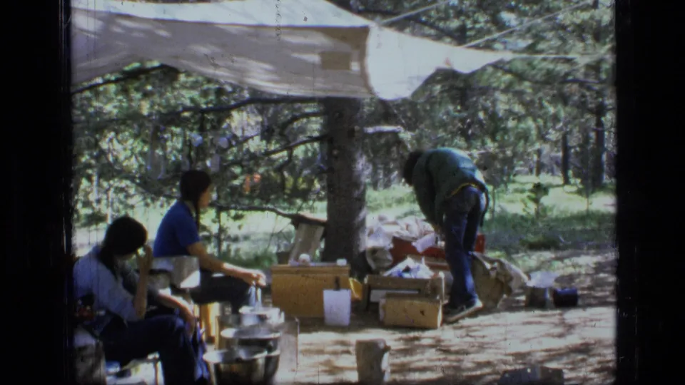 1973 Montana wilderness camping frame with seated figures, cooking pots, supply boxes, and a person sorting gear beneath a tarp.