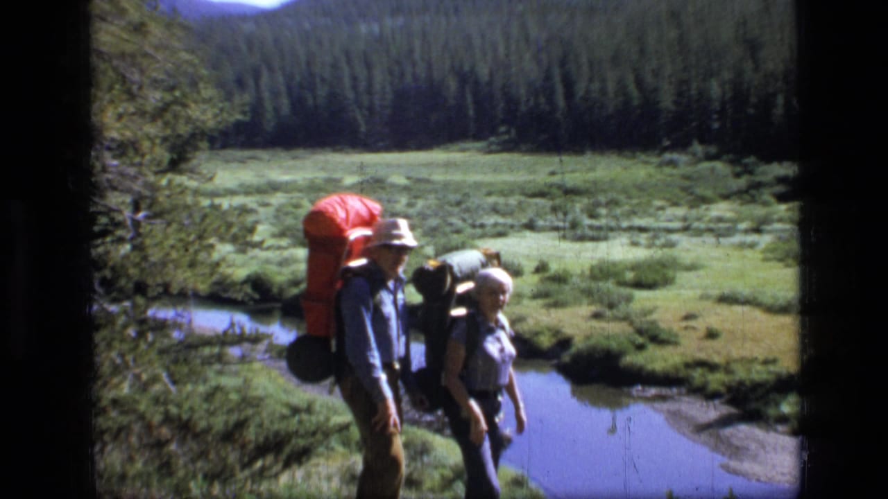 Two adult hikers in Scapegoat Wilderness, Montana, walk with large packs beside a meadow stream in 1973 home-movie footage.