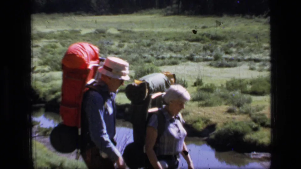 Closer 1973 view of two adult backpackers walking through Scapegoat Wilderness meadow terrain beside water.