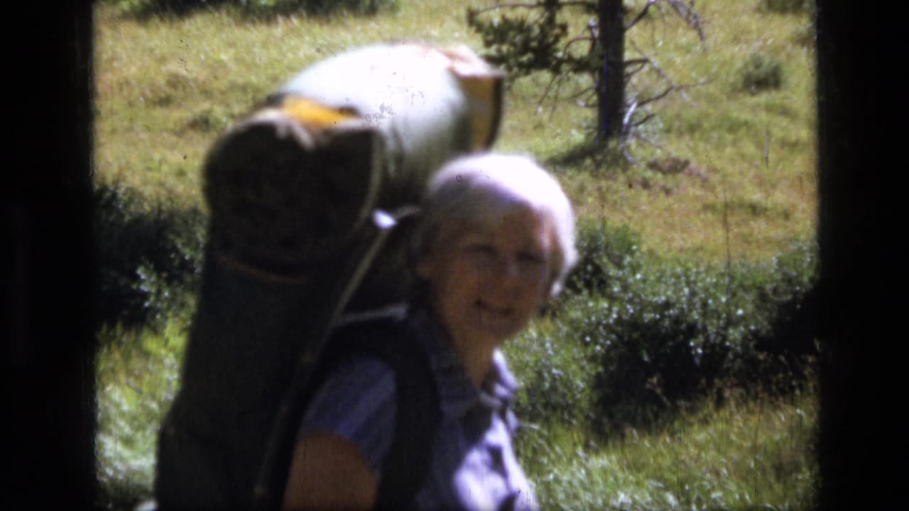1973 Montana home-movie frame of an adult woman with short white hair carrying a large backpack on a grassy trail.