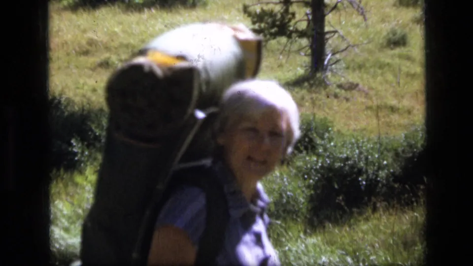1973 Montana home-movie frame of an adult woman with short white hair carrying a large backpack on a grassy trail.
